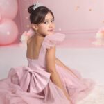 A young female model in a pink dress with a big bow, sitting and looking back.