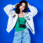 Teen female model posing confidently in a white jacket and green top against a blue background.