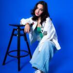 Teen model leaning on a black stool in a relaxed pose, wearing a graphic jacket.