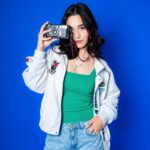 Model holding a vintage camera to her eye while posing against a vibrant blue background.