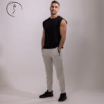 Full-length shot of a male model in Kuwait wearing a black sleeveless shirt, light grey joggers, and black sneakers, hands in pockets.