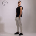 Full-length side profile of a male model in Kuwait smiling, wearing a black sleeveless shirt, light grey joggers, and black sneakers, hands in pockets.