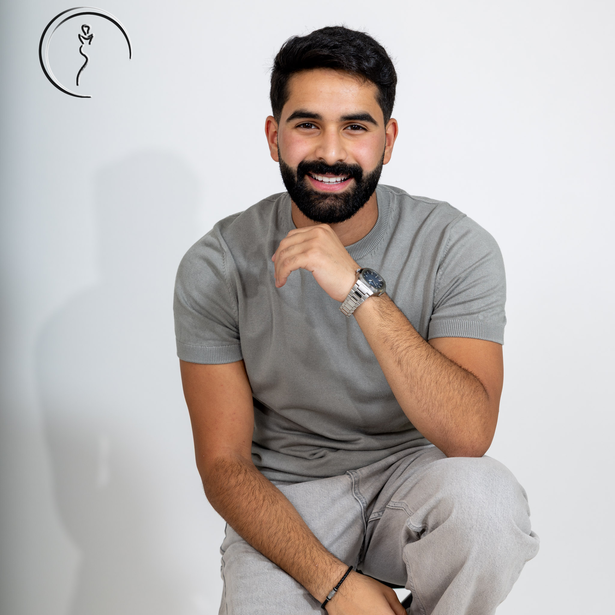 Man with a beard smiling, wearing a grey short-sleeved sweater and light grey jeans, with his hand on his chin.