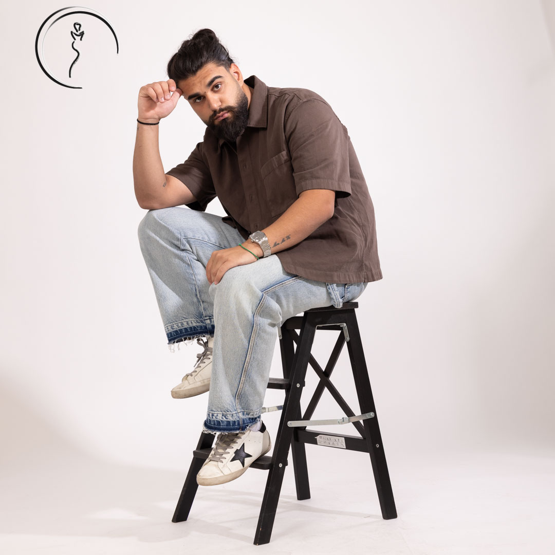 Male model with a beard and man bun, sitting on a stool, wearing a brown shirt and light wash jeans, with a thoughtful expression.