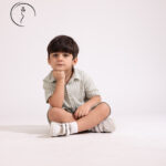 A young boy in a green checkered shirt and shorts sits cross-legged, smiling broadly with his hands on his cheeks.