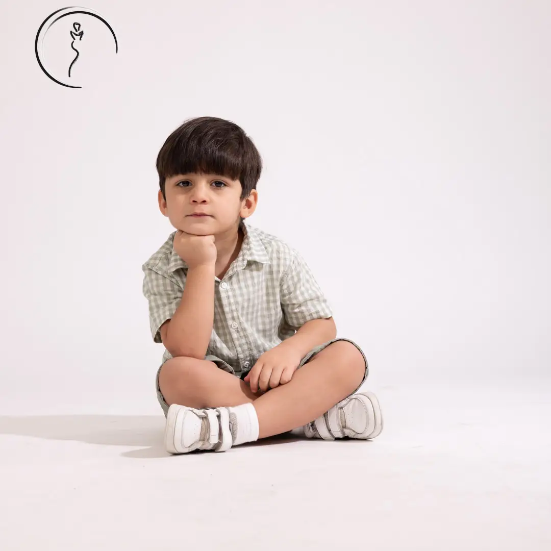 A young boy in a green checkered shirt and shorts sits cross-legged, smiling broadly with his hands on his cheeks.