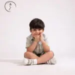 A young boy in a green checkered shirt and shorts sits cross-legged, resting his chin on his fist.