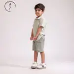 A young boy in a light green checkered shirt and shorts stands facing left, with his hands clasped in front of him.