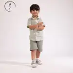 A young boy in a green checkered shirt and shorts smiles, clapping his hands together.