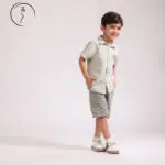 A young boy in a green checkered shirt and shorts stands smiling, looking to his right with his hands in his pockets.