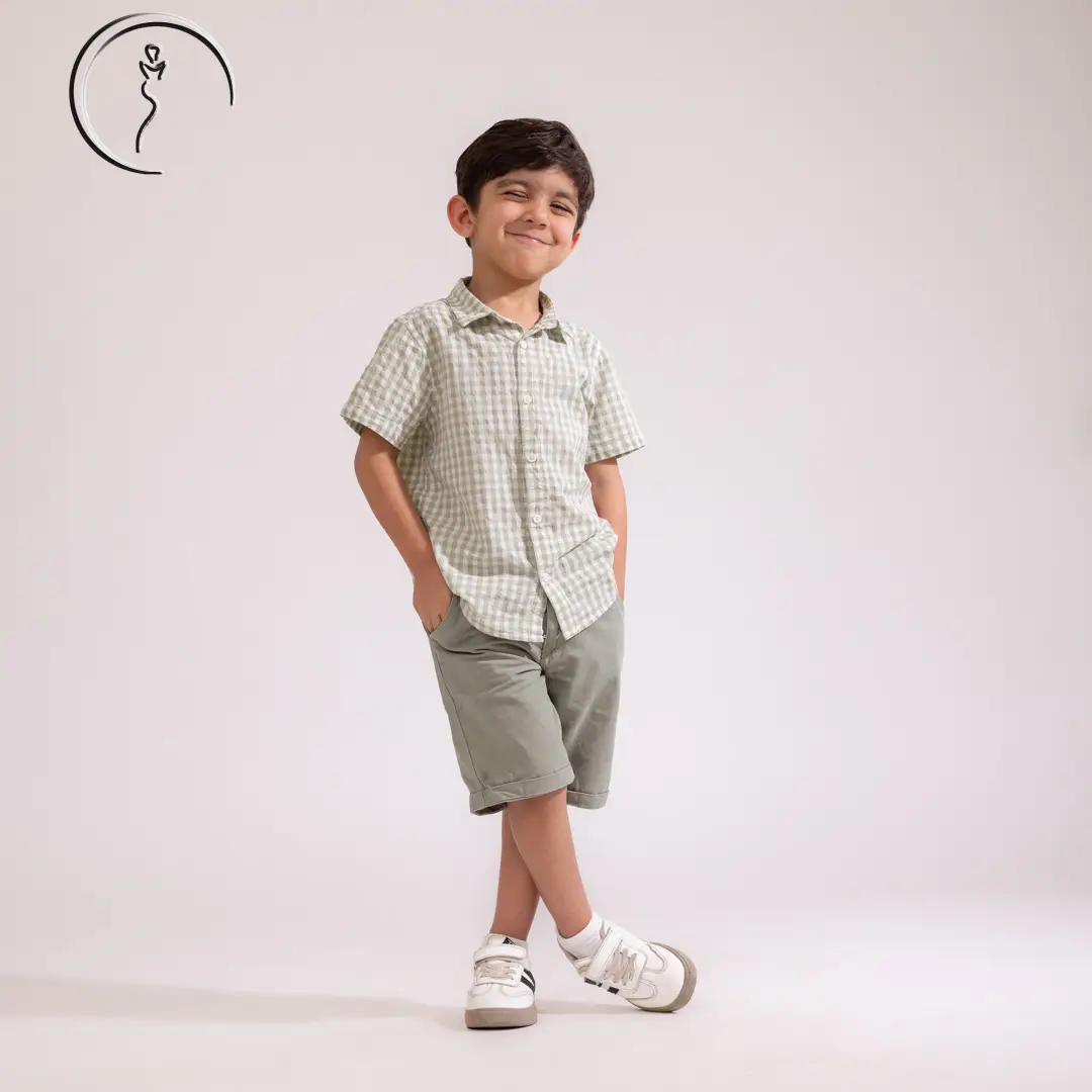 A young boy in a light green checkered short-sleeve shirt and matching shorts smiles widely with his hands in his pockets, his legs crossed.