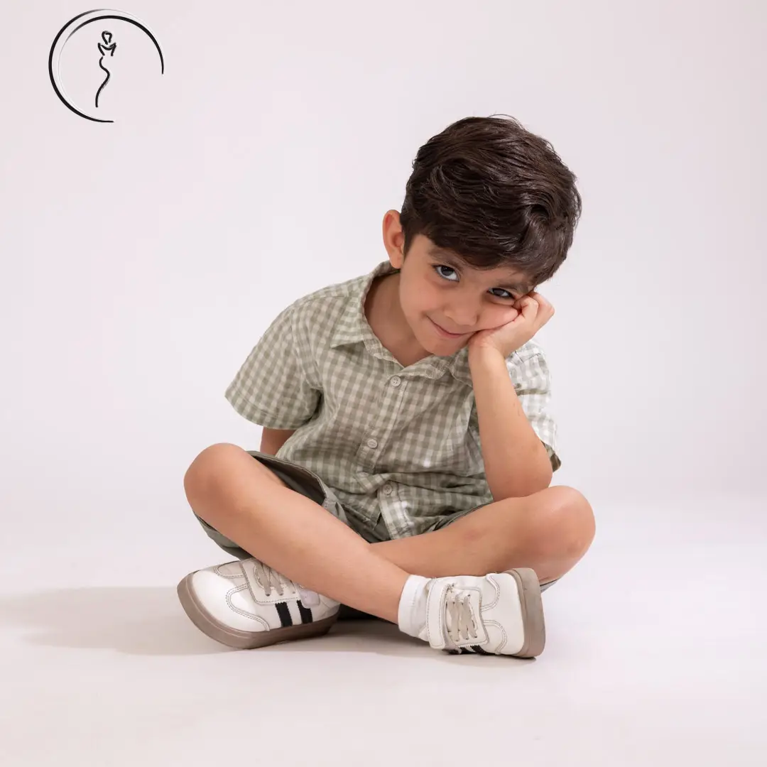 A young boy in a green checkered shirt and shorts sits cross-legged, resting his chin on his hand and looking thoughtfully at the camera.