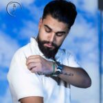 Male model with a beard and man bun, wearing an open brown short-sleeve shirt over a white t-shirt, hand on collar.