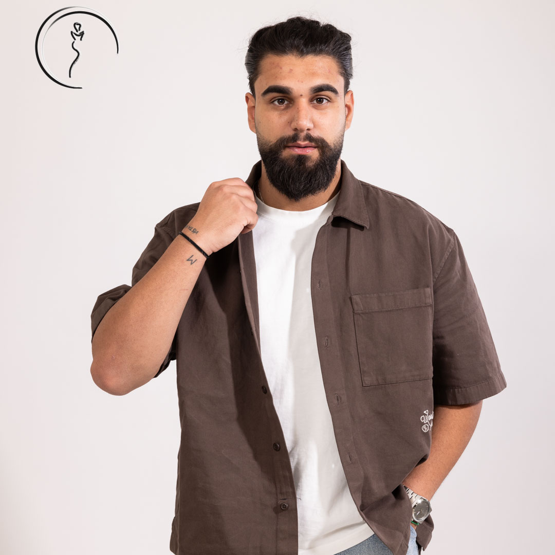 Close-up of a male model with a beard and man bun, looking down at his watch, wearing a white ribbed polo shirt.
