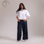 A full-body shot of a smiling woman posing in a white button-down shirt with wide, voluminous sleeves and high-waisted, dark-wash wide-leg jeans.