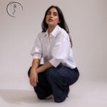 A woman crouching down and posing in a white button-down shirt with full sleeves and high-waisted, wide-leg denim jeans.