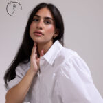 A close-up portrait of a woman with long dark hair, wearing a white shirt and gold earrings, looking directly at the camera. She has one hand gently touching her neck.