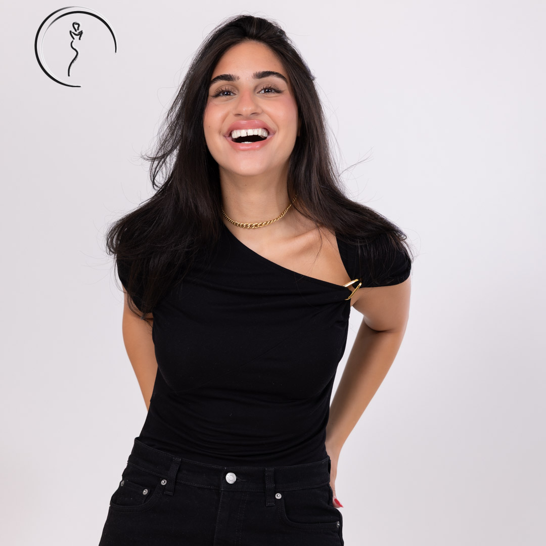 A candid, joyful shot of a woman with a big smile, wearing a black one-shoulder top with a gold chain accent.