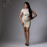 A full-body shot of an Indian model posing in a form-fitting, off-white ruched mini dress with a draped neckline and sparkling black heels.
