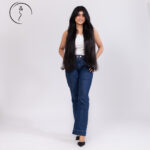A full-body shot of a smiling Indian model with long, dark hair wearing a white ribbed tank top and dark blue flared jeans with black heels.