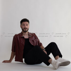 A male model in a red button-down shirt and black pants sits on the floor, smiling slightly at the camera.