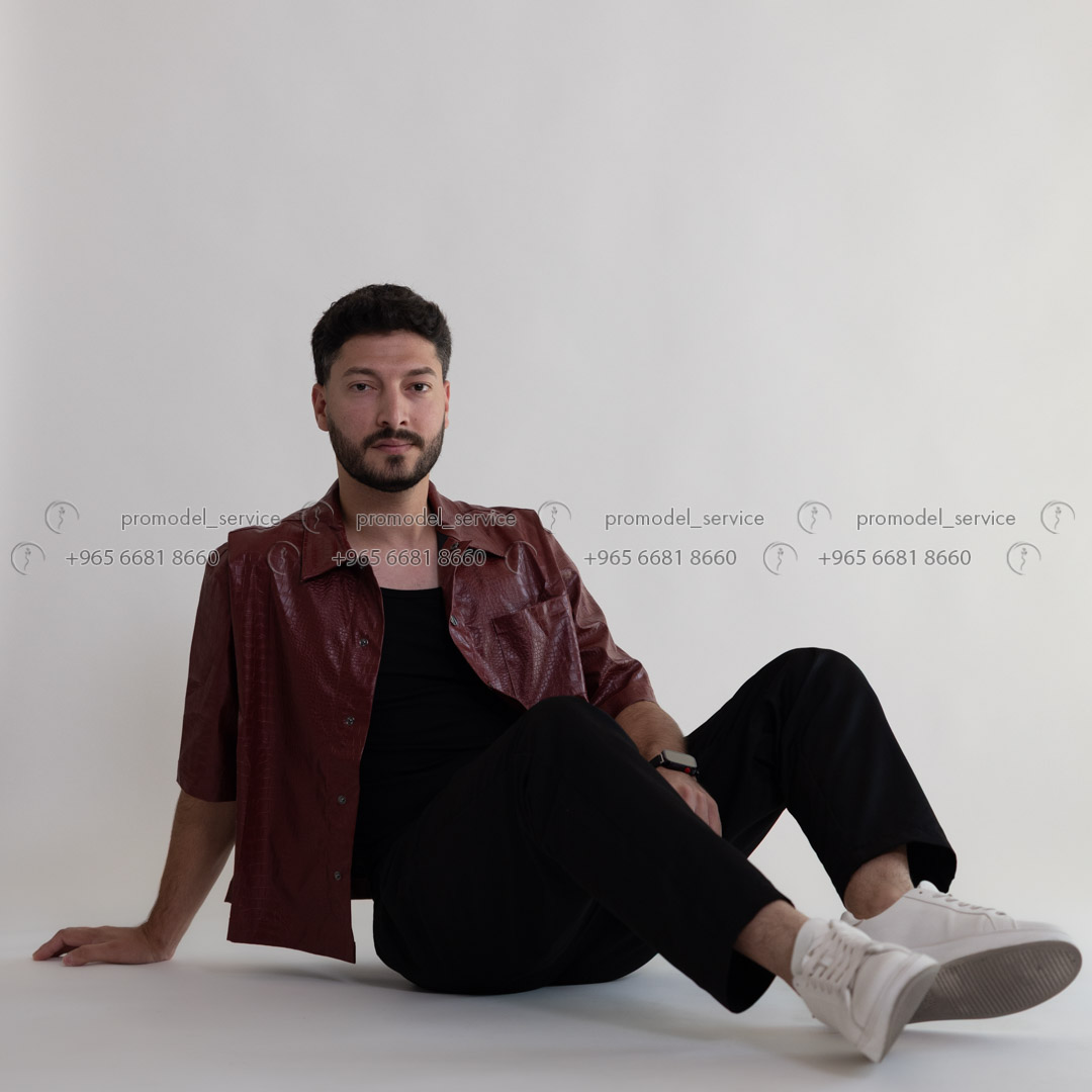 A male model in a red button-down shirt and black pants sits on the floor, smiling slightly at the camera.