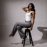 Female model on stool, white vest, gray jeans, high heels, leg up.
