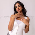 Close-up of female model, white vest, gold jewelry, hands near face.