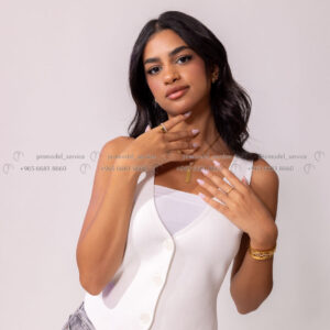 Close-up of female model, white vest, gold jewelry, hands near face.
