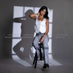 Female model sitting on stool, white vest, gray jeans, window shadow.