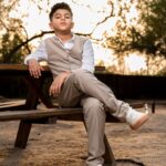 : A young boy in a tan vest and pants sits on a wooden picnic table, smiling.