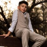A young boy in a tan vest and pants sits on a wooden picnic table, smiling at the camera.