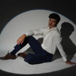 A male model with a mustache and curly dark hair wearing a white shirt, navy pants, and brown dress shoes, posing on the floor.