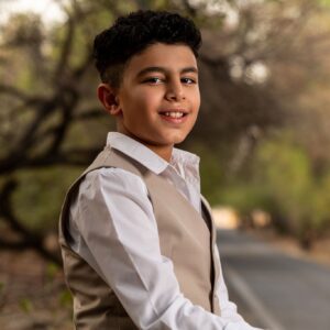 A young boy in a tan vest and pants sits on a wooden picnic table, looking at the camera.