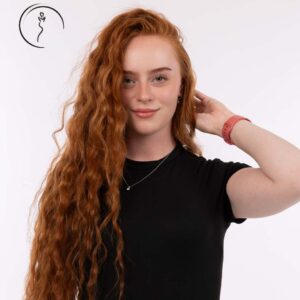 A red-haired woman smiling at the camera, with her hand on her hair.