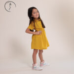 Female kid model smiling in a bright yellow buttoned dress with sneakers.