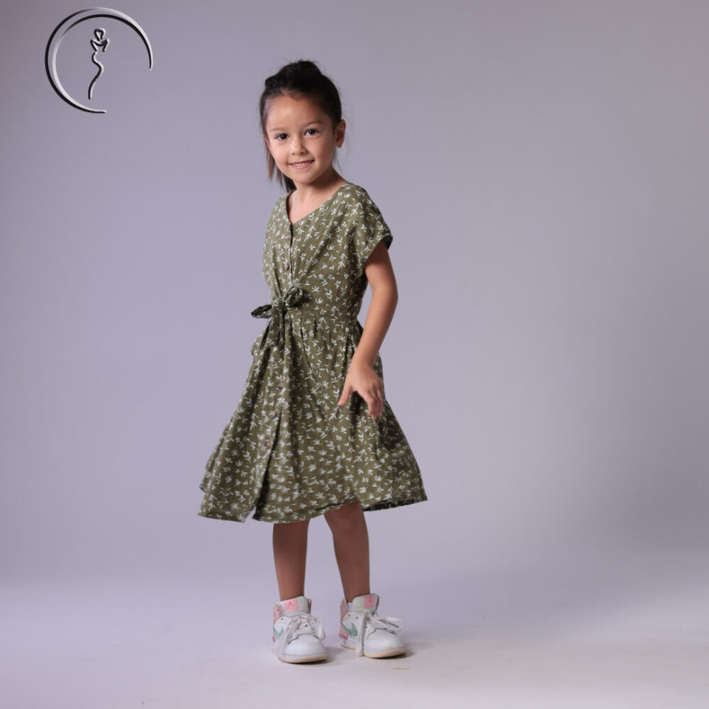 Child model in an olive green floral dress, standing naturally with a bright smile.