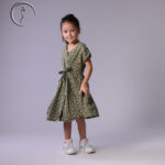 Child model in an olive green floral dress, standing naturally with a bright smile.