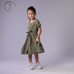 Child model in an olive green floral dress, standing naturally with a bright smile.
